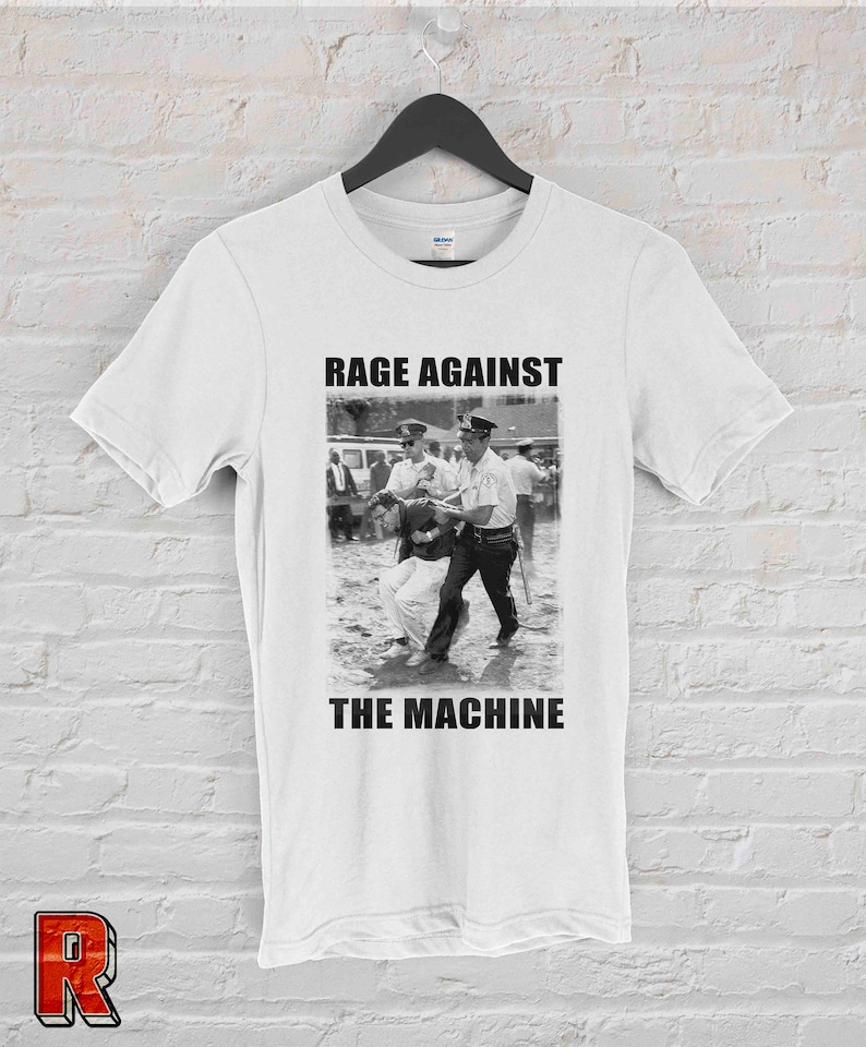 Op de afbeelding: Wit T-shirt met een zwart-wit afbeelding van een protestsc&egrave;ne en de tekst "RAGE AGAINST THE MACHINE". De afbeelding toont een persoon die door politieagenten wordt ge&euml;scorteerd. Het T-shirt hangt aan een zwarte hanger.