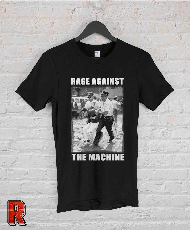 Op de afbeelding: Zwart T-shirt met een zwart-wit protestbeeld en de witte tekst "RAGE AGAINST THE MACHINE". Het shirt hangt aan een zwarte hanger tegen een witte bakstenen muur.