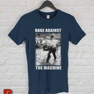 Op de afbeelding: Marineblauw T-shirt met een zwart-wit protestbeeld en de witte tekst "RAGE AGAINST THE MACHINE". Het T-shirt hangt aan een zwarte hanger tegen een witte bakstenen muur.