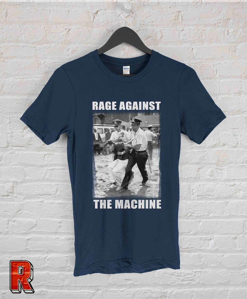 Op de afbeelding: Marineblauw T-shirt met een zwart-wit protestbeeld en de witte tekst "RAGE AGAINST THE MACHINE". Het T-shirt hangt aan een zwarte hanger tegen een witte bakstenen muur.