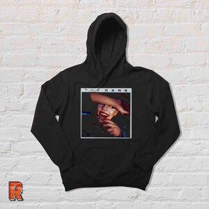 May include: Black hooded sweatshirt with a graphic print of the band The Cars album cover. The album cover features a woman with red lipstick and a hand covering her face.