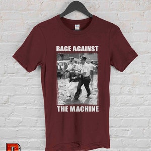 Op de afbeelding: Bordeauxrood t-shirt met een zwart-wit afbeelding van een protestsc&egrave;ne en de tekst "RAGE AGAINST THE MACHINE" in witte blokletters. Het shirt hangt aan een zwarte hanger tegen een witte bakstenen muur.