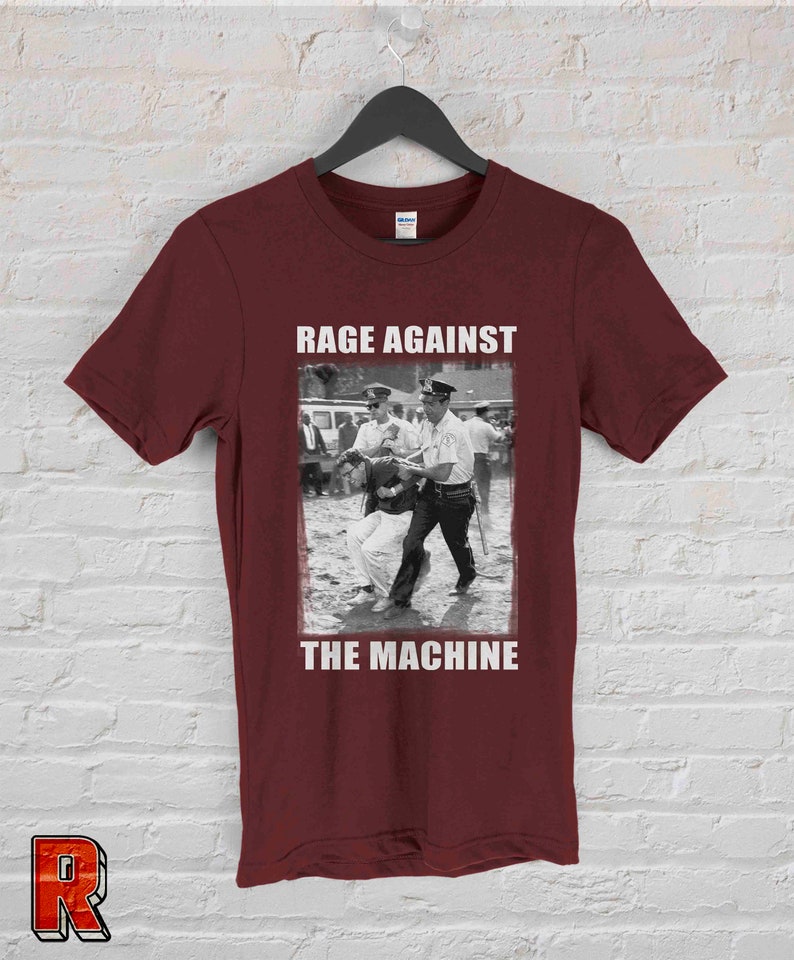 Op de afbeelding: Bordeauxrood t-shirt met een zwart-wit afbeelding van een protestsc&egrave;ne en de tekst "RAGE AGAINST THE MACHINE" in witte blokletters. Het shirt hangt aan een zwarte hanger tegen een witte bakstenen muur.
