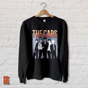 May include: Black sweatshirt with a vintage-style graphic of the band "The Cars" in orange and yellow text. The band members are pictured in black and white.