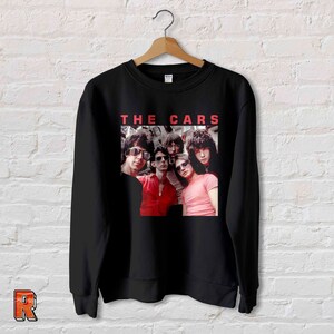 May include: Black sweatshirt featuring a band photo of The Cars with the band's name in red block letters above the image. The band members are wearing sunglasses and various colored shirts.