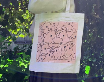 May include: A white tote bag with a pink rectangular design featuring a pattern of cartoon bunnies. The bunnies are in various poses and are outlined in black. The bag is held up against a background of green foliage.