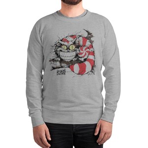 May include: Gray sweatshirt featuring a whimsical illustration of the Cheshire Cat from Alice in Wonderland. The cat has a large grin, yellow eyes, and a striped red and white tail. The design is a detailed black and white drawing with red accents.