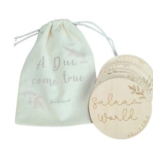 May include: A cream-coloured drawstring bag with the text "A Dua come true" and a set of wooden round discs. The discs are engraved with the words "Salaam World" and a leaf design. The bag and discs are light in colour.