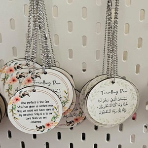 May include: A selection of circular wooden pendants suspended on silver ball chains. Each pendant displays a different design, including floral motifs, religious text, and the words "Travelling Dan". The pendants are shown against a white backdrop.