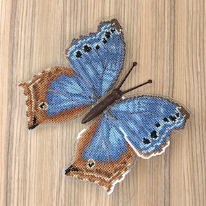 May include: A blue and brown butterfly with a beaded design is displayed on a wood background. The butterfly is made of fabric and has a three-dimensional appearance.