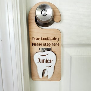 Peut inclure: Panneau de porte en bois avec le texte "Dear toothfairy Please stop here" et une poche en forme de dent avec le mot "Junior". Le panneau est accroché à une porte blanche. Un billet de cinq dollars est visible dans la poche.
