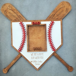 May include: Wooden baseball-themed photo frame with crossed bats. The frame is shaped like a home plate, with red stitching details and the word "DAD" in red. Text includes "WE HIT A HOME RUN WITH YOU" and names.