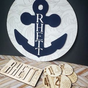 Puede incluir: Un letrero de madera para hitos de bebé con temática náutica con un ancla azul y el nombre "Rhett" escrito verticalmente. El letrero está rodeado de un fondo blanco con un patrón de olas. También hay círculos de madera separados con números y la palabra "meses" en ellos.
