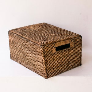 Op de afbeelding: Een rechthoekige opbergmand van geweven rotan met een deksel. De mand is in natuurlijke tinten met donkerbruine accenten. Hij heeft handvatten aan de zijkanten en is ontworpen voor opslag.
