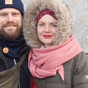 May include: A couple wearing winter jackets and hats. The woman is wearing a pink scarf and a red and white corduroy fanny pack. The man is wearing a navy blue beanie and a green corduroy fanny pack.