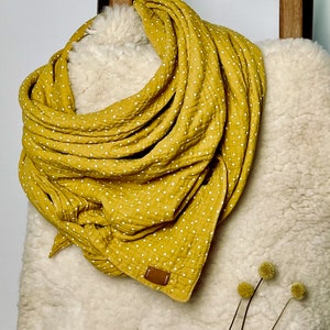 May include: Mustard yellow scarf with white polka dots, draped over a cream-colored, textured surface. The scarf has a small brown leather tag. The background includes a wooden structure and dried yellow flowers.