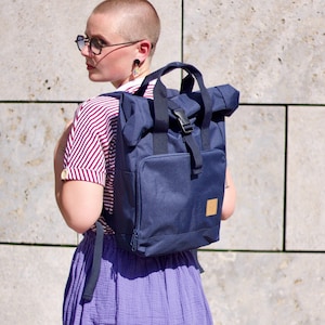 Könnte beinhalten: Ein marineblauer Rucksack mit Rollverschluss, Vordertasche und Schnallenverschluss. Der Rucksack hat ein kleines, quadratisches, hellbraunes Patch. Die Person trägt ein gestreiftes Hemd, lila Shorts und eine Brille.