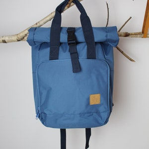 Op de afbeelding: Een blauwe roll-top rugzak met een gespsluiting en een bruin leren patch. De rugzak heeft verstelbare banden en een ritsvak.