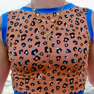 Può includere: Un top marrone senza maniche con stampa leopardata. Il top ha un bordo blu intorno al collo e all'orlo inferiore. La modella indossa una collana a catena dorata a più strati.