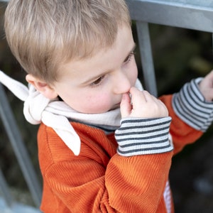 May include: A young boy wearing a bright orange corduroy sweater with white stripes on the sleeves and a white bandana around his neck. He is holding onto a metal railing with one hand and picking his nose with the other.