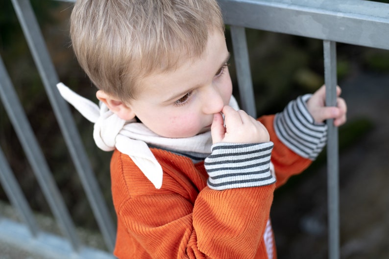 May include: A young boy wearing a bright orange corduroy sweater with white stripes on the sleeves and a white bandana around his neck. He is holding onto a metal railing with one hand and picking his nose with the other.