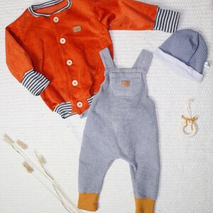 May include: A set of baby clothes in orange, gray, and white. The set includes a corduroy jacket with striped cuffs, a pair of gray corduroy overalls with orange cuffs, and a white and gray polka dot hat.