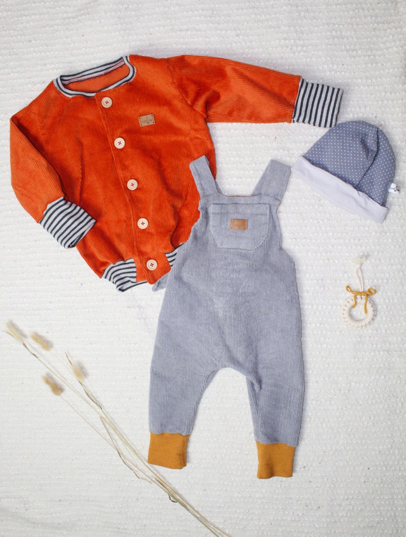 May include: A set of baby clothes in orange, gray, and white. The set includes a corduroy jacket with striped cuffs, a pair of gray corduroy overalls with orange cuffs, and a white and gray polka dot hat.