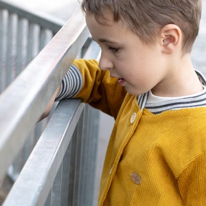 May include: A young boy wearing a yellow corduroy jacket with a striped collar and a wooden button. The jacket has a small wooden patch on the front.