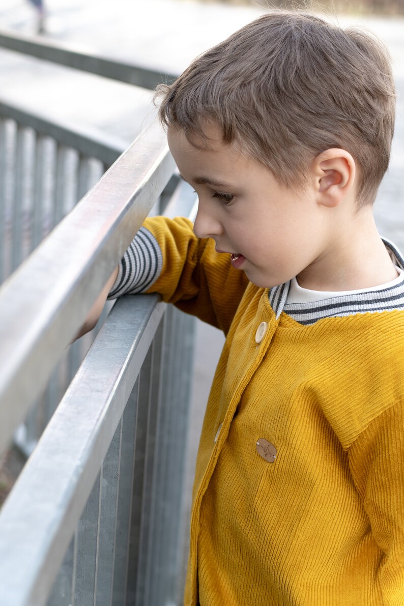 May include: A young boy wearing a yellow corduroy jacket with a striped collar and a wooden button. The jacket has a small wooden patch on the front.