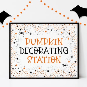May include: A white sign with black and orange confetti and the text "Pumpkin Decorating Station" in black. There are two black spiders on the sign.