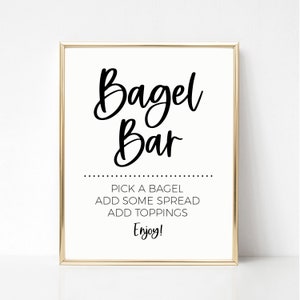 Könnte beinhalten: Ein weißes Schild mit goldenem Rahmen, auf dem in schwarzer Kursivschrift "Bagel Bar" steht. Unter dem Titel steht: "Wählen Sie einen Bagel, fügen Sie etwas Streichwurst hinzu, fügen Sie Beläge hinzu, Genießen Sie!"