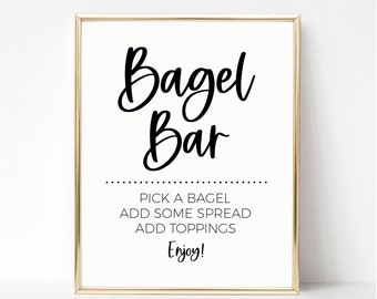 May include: A white sign with a gold frame that says "Bagel Bar" in black cursive font. Below the title, it says "Pick a bagel, add some spread, add toppings, Enjoy!"