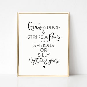 Puede incluir: Un cartel blanco con marco dorado, con el texto "Grab a prop & strike a pose... serious or silly Anything goes!" en fuente cursiva negra.