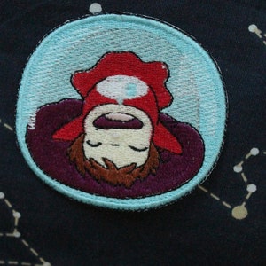 May include: A blue and white embroidered patch featuring a cartoon character with brown hair, red clothing, and a white face. The character is lying on their back inside a bubble.