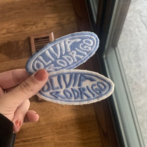 May include: Two blue and white oval embroidered patches with the text "Olivia Rodrigo" in white.