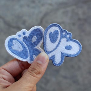 Op de afbeelding: Twee geborduurde patches in de vorm van een vlinder met een tweekleurig ontwerp in lichtblauw en wit. Elke patch toont een gestileerd vleugelpatroon met hartvormige uitsparingen en afgeronde randen, afgewerkt met een dikke witte stikrand.