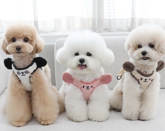 May include: Three small dogs wearing adorable knitted dog harnesses with bear faces. The harnesses are in pink, black and white, and brown. The dogs are sitting on a white couch.