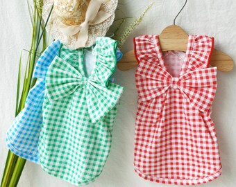 May include: Two gingham doll dresses, one green and one red, with ruffled trim and bow accents. The dresses are on hangers and a straw hat with lace trim is in the background.