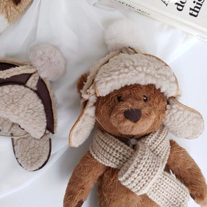 Puede incluir: Un oso de peluche marrón que lleva un gorro de piel sintética de color crema con orejeras y una bufanda de punto de color crema. El oso tiene la nariz marrón y los ojos negros.