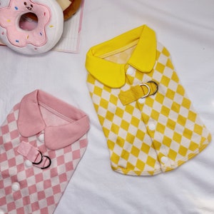 May include: Two dog shirts with a checkered pattern. One shirt is yellow with a yellow collar and the other is pink with a pink collar. Both shirts have a silver buckle and a button closure.