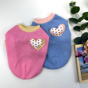 May include: Two pet sweaters, one pink with a yellow ribbed collar and one blue with a light pink ribbed collar. Each sweater features a heart-shaped patch on the chest, consisting of a white heart with small red hearts printed on it.