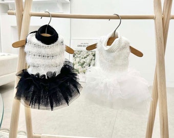 Può includere: Due vestiti per animali domestici appesi a un supporto in legno. L'abito a sinistra ha un collo alto nero, un corpetto bianco soffice e una gonna in tulle nero a strati. L'abito a destra è interamente bianco con collo alto e gonna in tulle.
