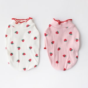 May include: Two pet shirts with a strawberry print. One shirt is white with red trim and the other is pink with red trim.