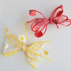 May include: Two hair bows, one yellow with white daisy embroidery and one red with white strawberry embroidery.