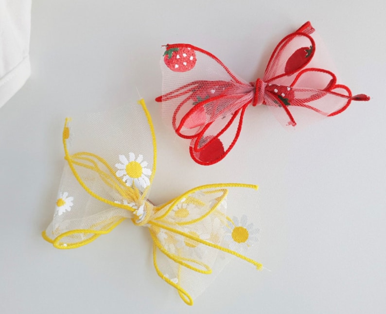 May include: Two hair bows, one yellow with white daisy embroidery and one red with white strawberry embroidery.