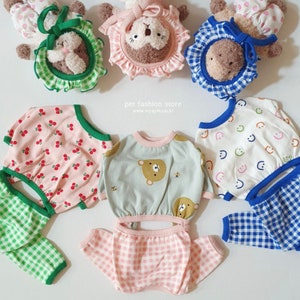 May include: A collection of pet clothing and accessories. The image features several outfits with various patterns, including cherries, gingham, and smiley faces. There are also plush animal headbands. The color palette includes pink, green, blue, and white. The text "pet fashion store" is visible.