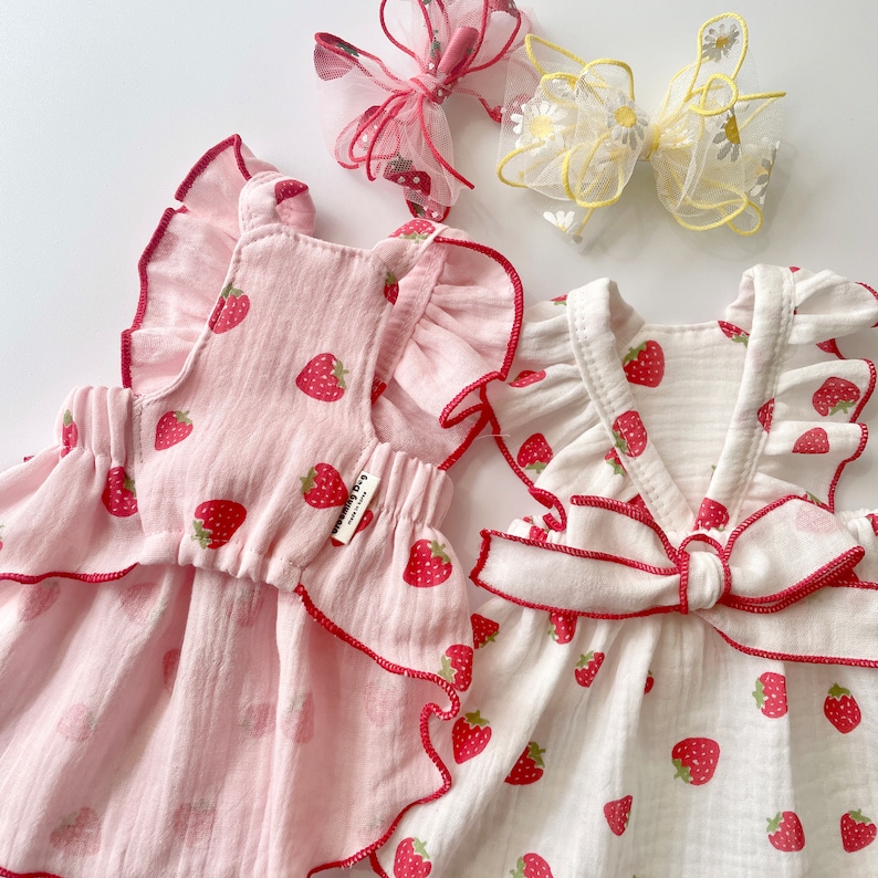 May include: Two white cotton baby dresses with strawberry print. One dress is pink with ruffled trim and the other is white with a bow.
