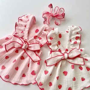 May include: Two white and pink sleeveless dresses with strawberry print and red ribbon bows. The dresses have a ruffled neckline and are made of a soft, lightweight fabric.