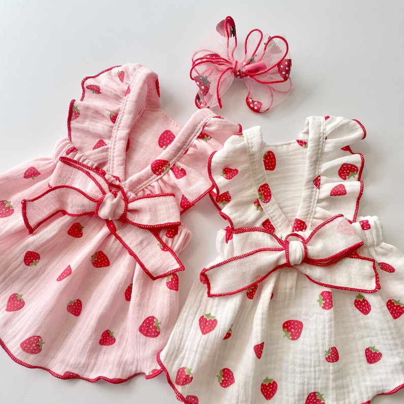 May include: Two white and pink sleeveless dresses with strawberry print and red ribbon bows. The dresses have a ruffled neckline and are made of a soft, lightweight fabric.