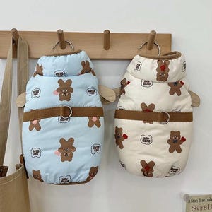 Peut inclure: Deux manteaux pour chien avec un imprimé d'ours brun. Un manteau est bleu clair avec une ceinture marron et l'autre est crème avec une ceinture marron.